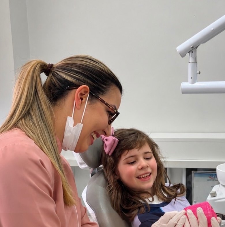 Dra. Karen Heiden sorrindo ao lado de uma menina sentada na cadeira do consultório, que segura o estojo cor-de-rosa do aparelho